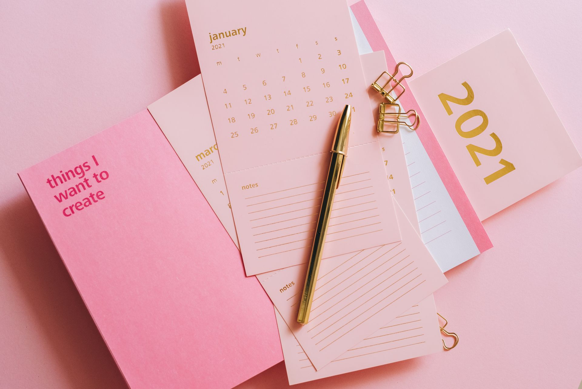 Elegant 2021 calendar setup with gold pen, clips, and planners on a pink background for organization inspiration.