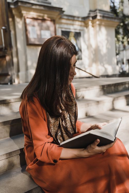Free Woman in a red dress reading a book while sitting on outdoor steps, enjoying a sunny day. Stock Photo