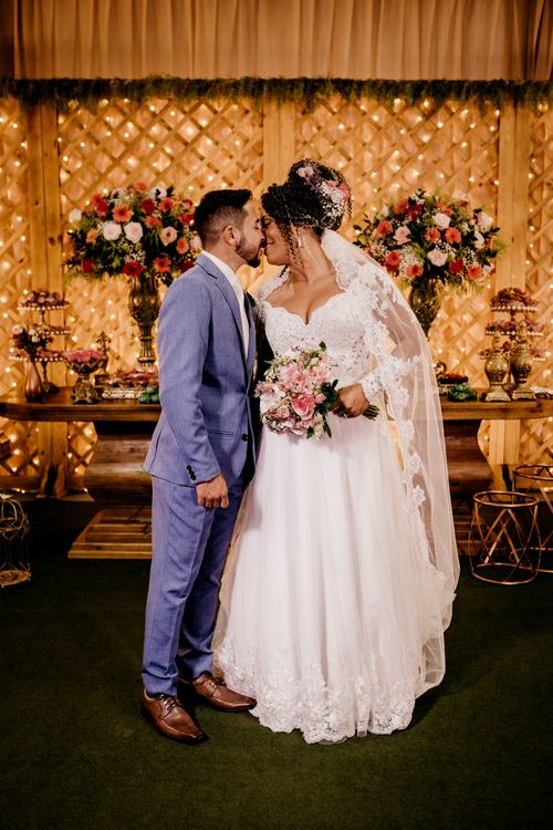 Free Young bride in wedding dress with bouquet of beautiful flowers standing near groom in stylish suit and kissing gently Stock Photo