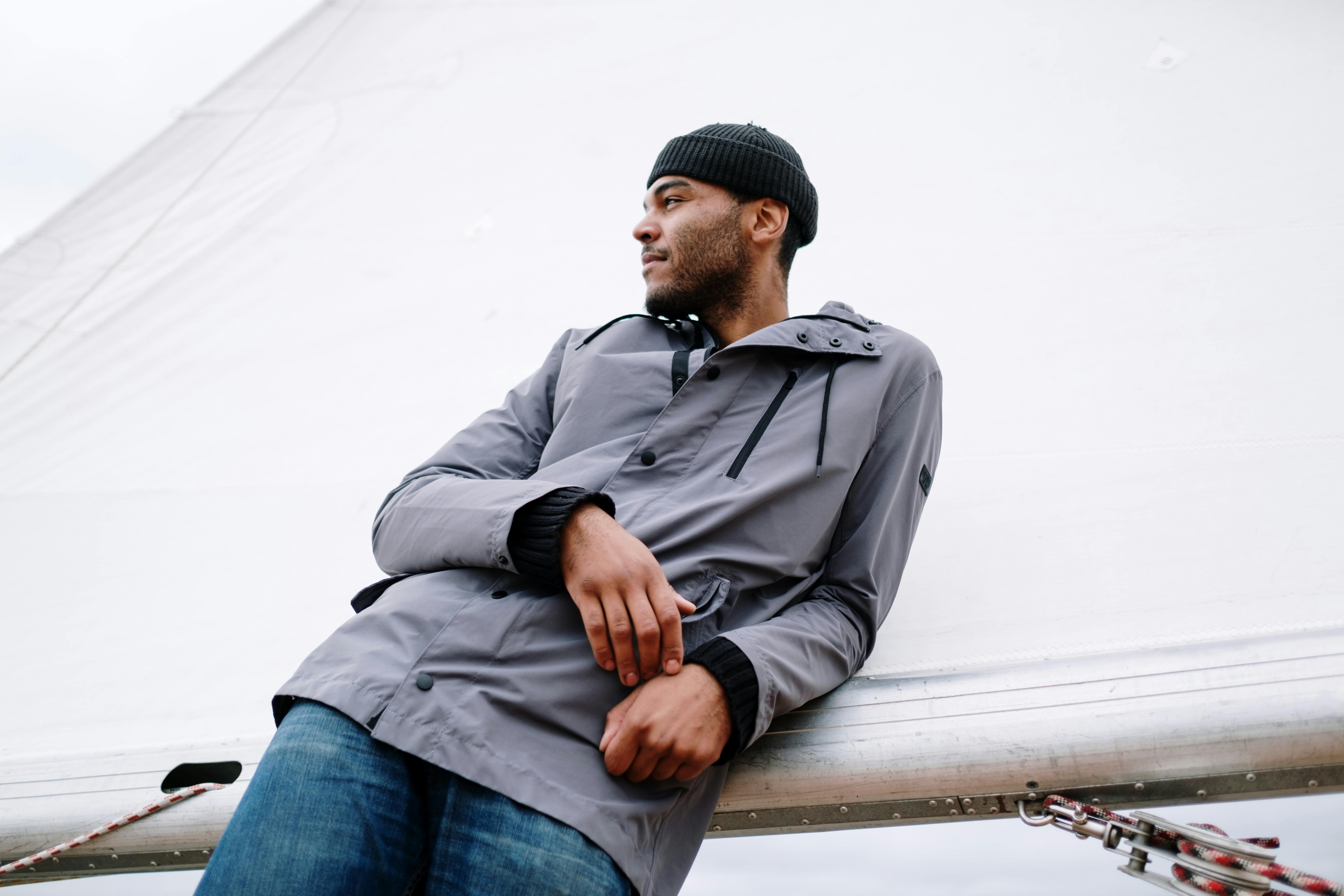 A man in a beanie and jacket leaning against a sail on a boat.