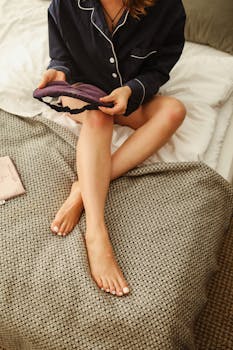 Relaxed woman in pajamas holding a sleep mask, sitting on a bed, embracing cozy comfort.