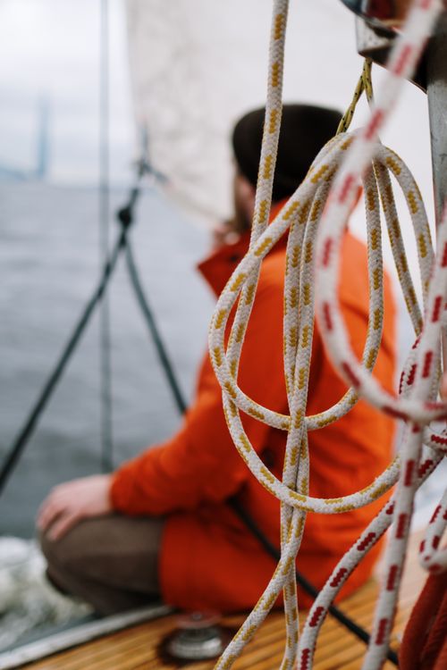 Free A man wearing an orange jacket enjoys a relaxing sail on a sailboat, focused on ropes and sea view. Stock Photo