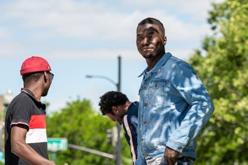Free Low angle of African American man in denim jacket looking at camera and standing near friends  on sunny street Stock Photo