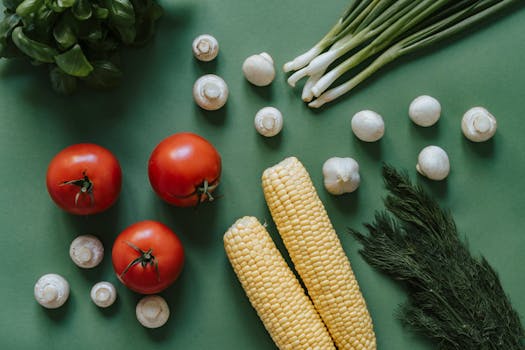 A top view of fresh vegetables including tomatoes, corn, and mushrooms on a green background.