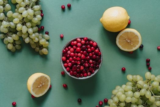 A vibrant flat lay of fresh cranberries in a bowl with green grapes and lemons on a green background.