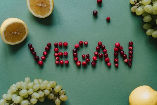 Vegan spelled out with cranberries, surrounded by lemons and grapes on a green surface.