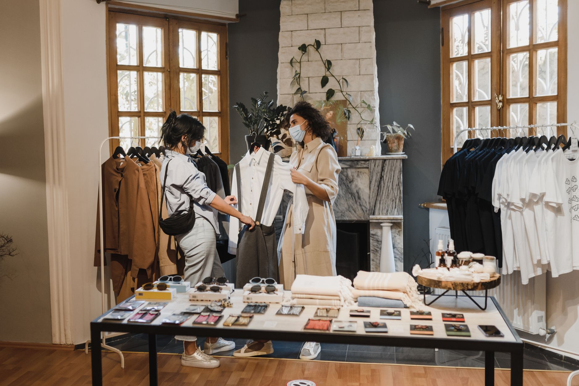 Two women wearing face masks shopping in a stylish boutique, browsing clothes on racks indoors.