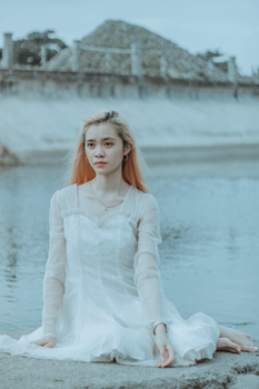 白天，一位头发有光、身着白衣的平静亚洲女士坐在平静水边的石面上，面对着有栅栏的混凝土结构
