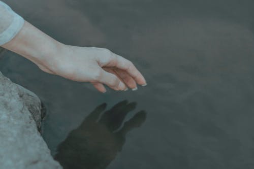 Crop anonymous female on boulder on coast near pond touching peaceful water with hand in daylight