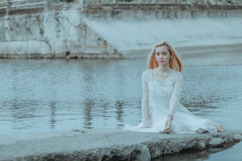 Free Calm young Asian female in elegant white dress sitting on stone surface near water in daytime Stock Photo