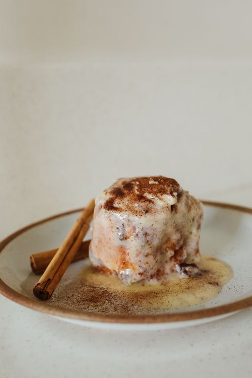 Free Close-up of a frosted cinnamon roll with cinnamon sticks on a plate. Perfect for dessert and food styling. Stock Photo