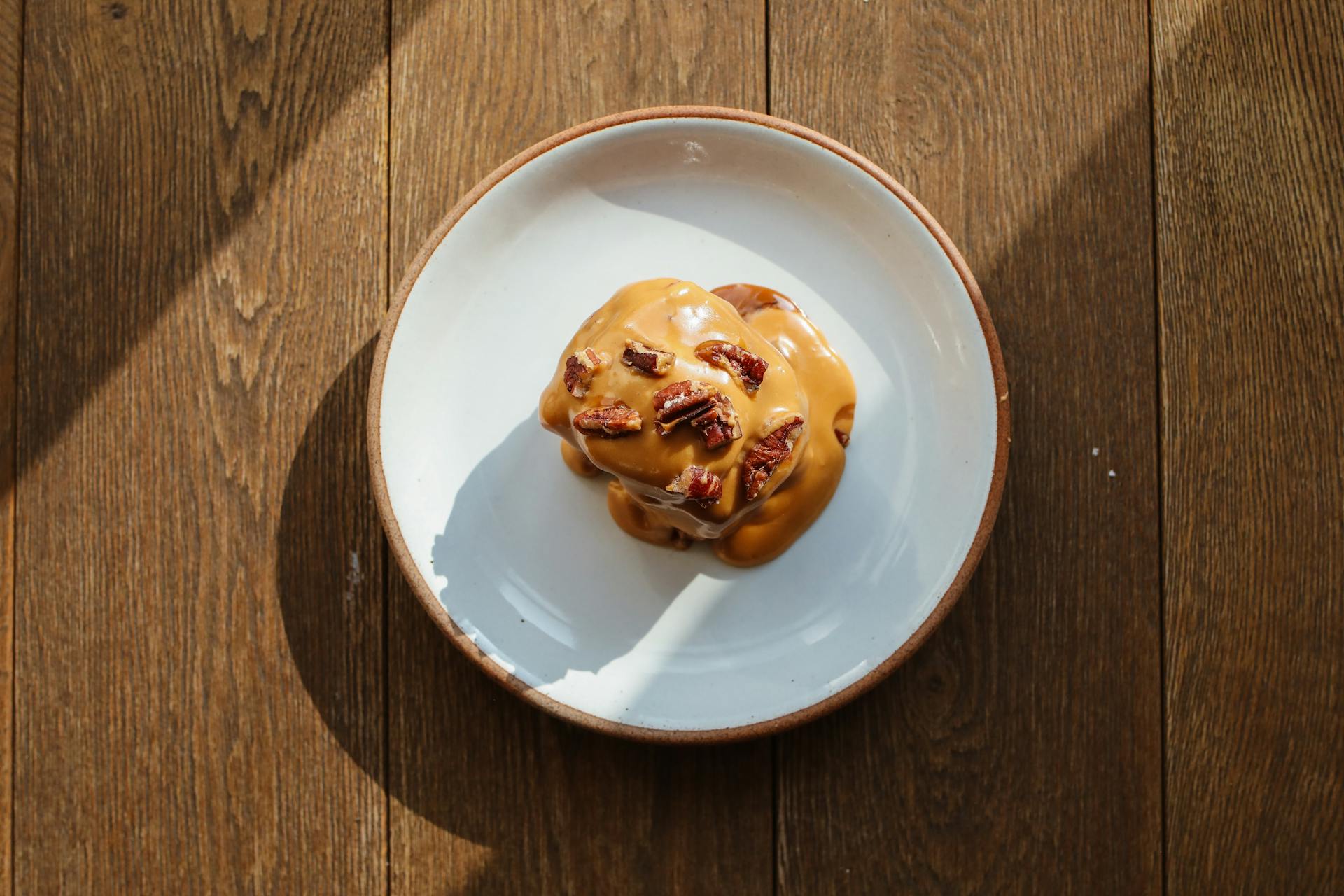 Top view of a sweet caramel-covered bun with pecans on a plate, sunlight on wooden table.