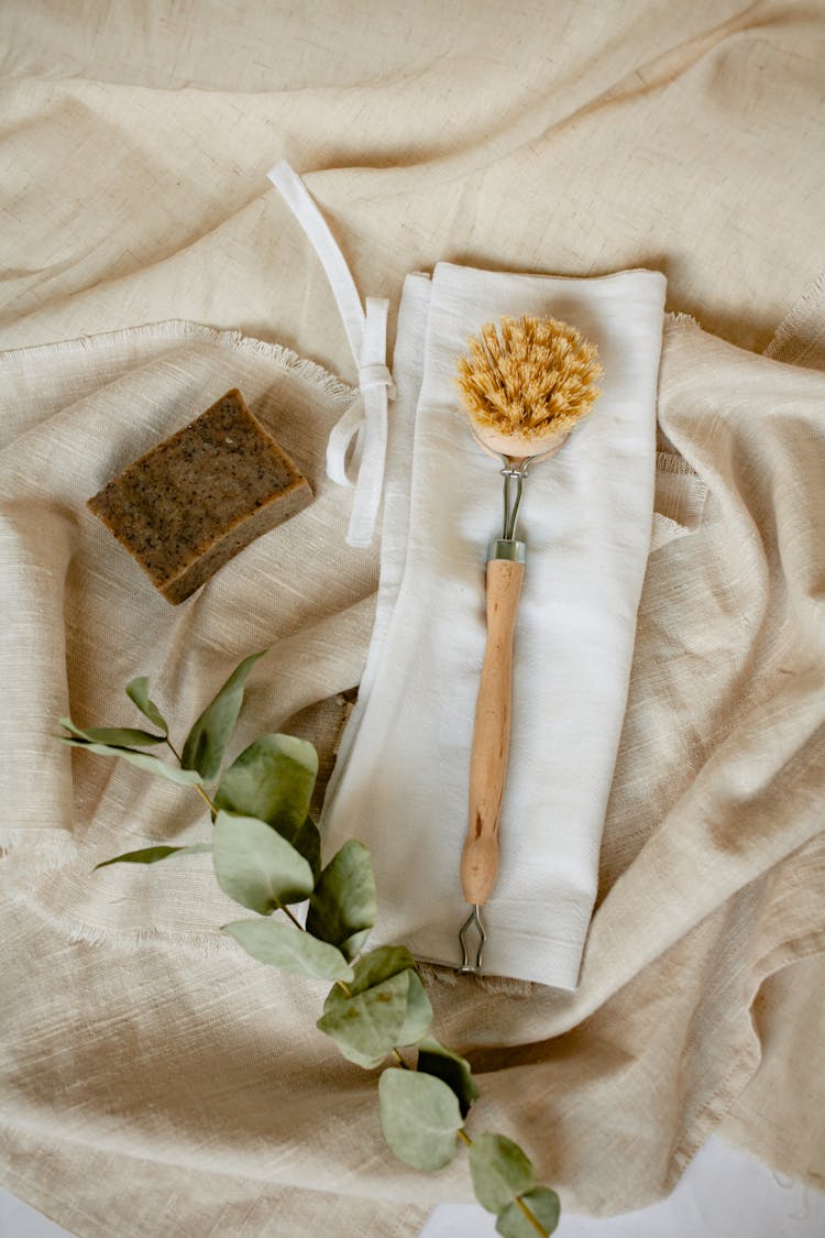 A Body Brush With Wooden Handle On A Folded Textile Beside A Bar Soap
