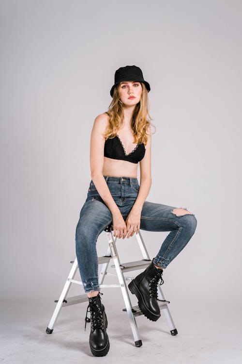 Free Full body of calm female with blond hair in hat and boots wearing black bra and ripped jeans sitting on metal ladder on white background and looking at camera Stock Photo