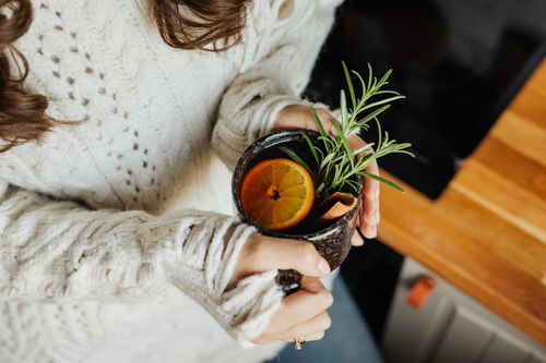 Free A woman in a cozy sweater holds a herbal tea with citrus and rosemary, evoking autumn warmth. Stock Photo