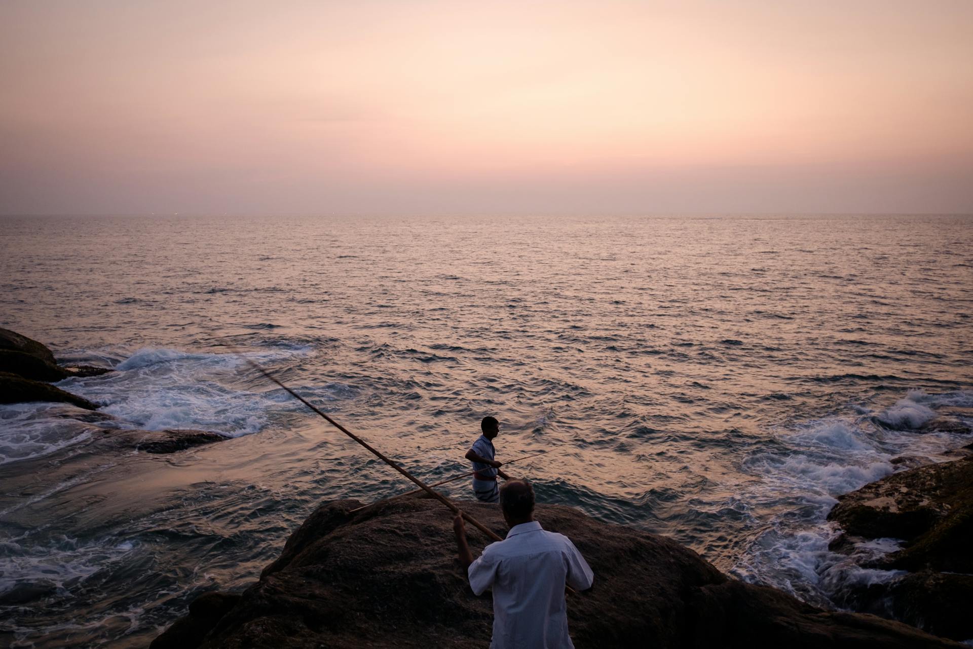 Two fishermen at sunset casting rods along the rocky Sri Lankan coast.