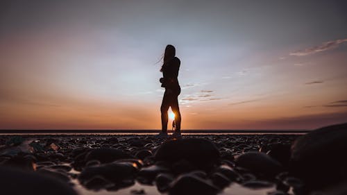 Side view silhouette of unrecognizable female traveler relaxing on ocean shore during low tide against sunset sky