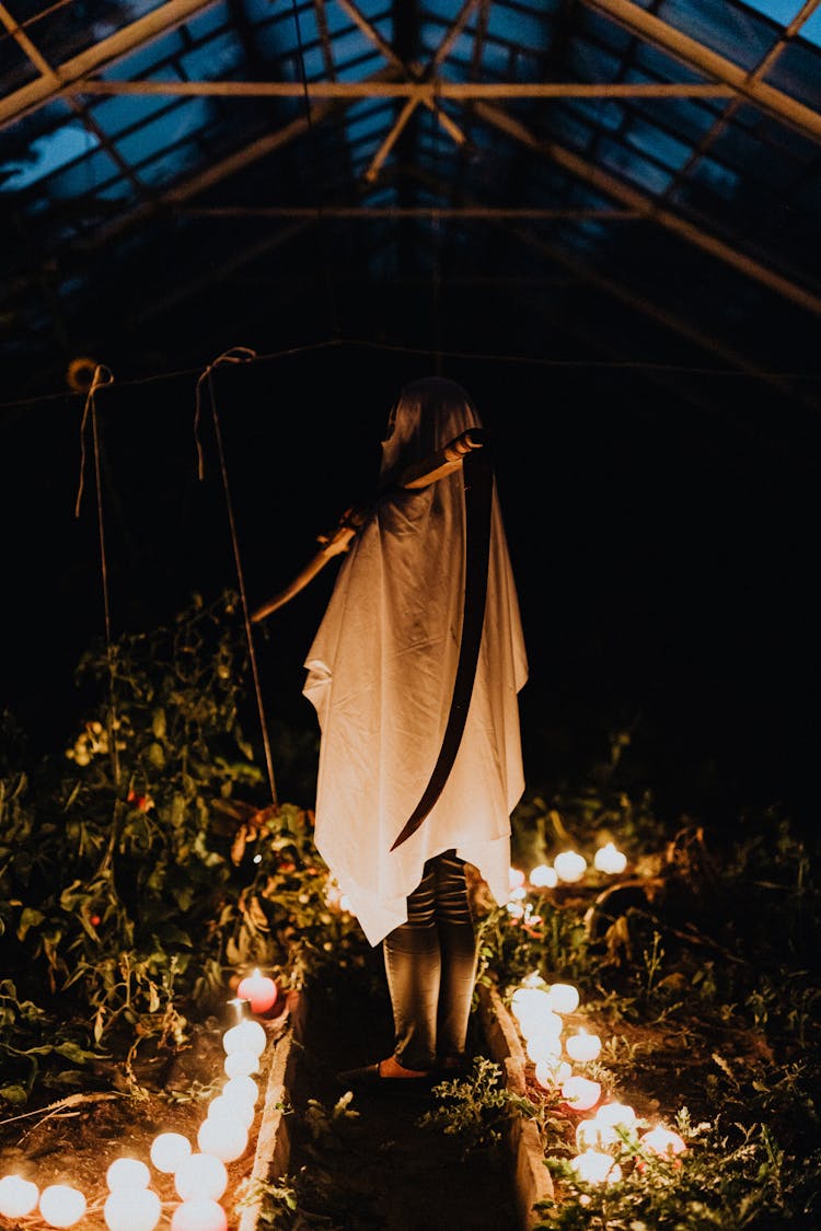 Person Wearing A White Sheet And Holding A Scythe Standing In A Greenhouse At Night 