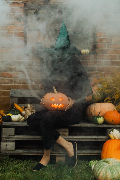 Free Person in Black Pants Sitting on Brown Wooden Bench With Jack O Lantern on His Lap Stock Photo