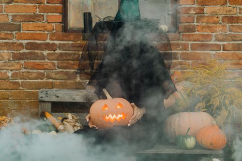 Free Mysterious witch holding a glowing Jack-O'-Lantern in a smoky, festive Halloween setting. Stock Photo
