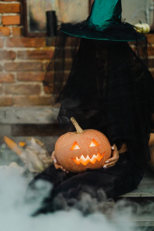 Free A witch in black attire holds a jack-o'-lantern against a brick wall on a foggy night. Stock Photo