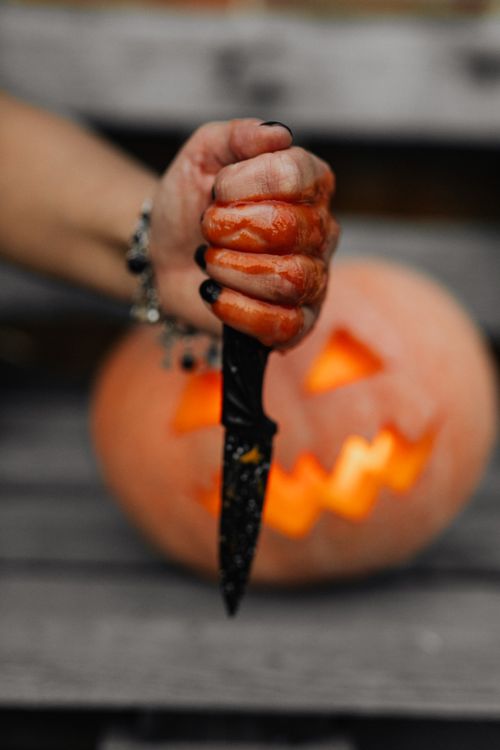 Free Close-up of a bloody hand holding a knife in front of a carved pumpkin, perfect for Halloween themes. Stock Photo