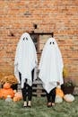 两个穿着幽灵服装的人站在质朴的砖墙前，周围环绕着南瓜，体现了万圣节精神
