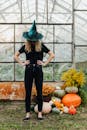 一名女巫打扮的女人站在布满蜘蛛网的温室里，周围堆满了南瓜