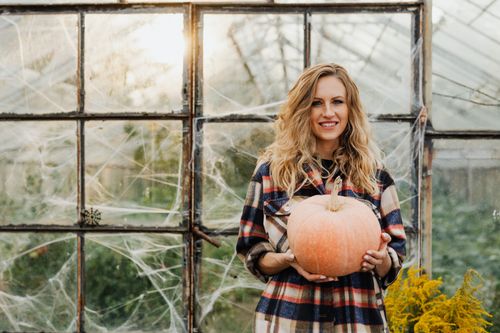 Free Smiling woman holding a pumpkin in a fall-themed greenhouse, ideal for autumn and holiday concepts. Stock Photo