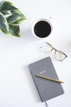 极简主义工作空间的顶视图显示，白色桌面上摆放着咖啡、笔记本和绿植