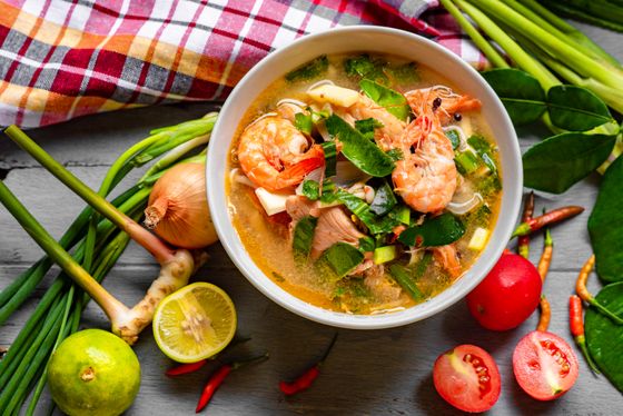 Bowl of clear hot and sour shrimp soup with greens, surrounded by lemongrass, limes and chilies