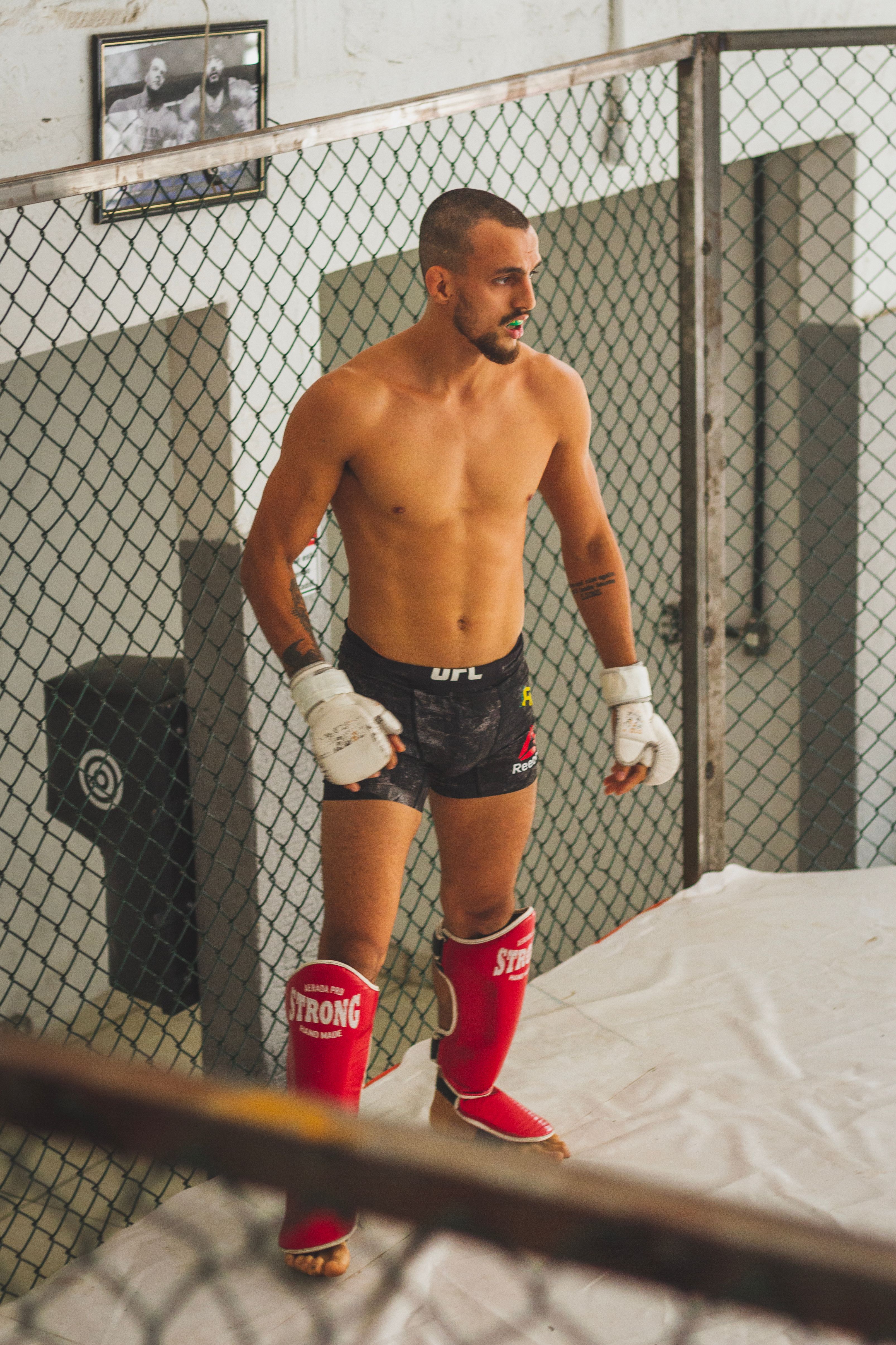 MMA fighter wearing shin guards and gloves preparing for practice in a training cage.