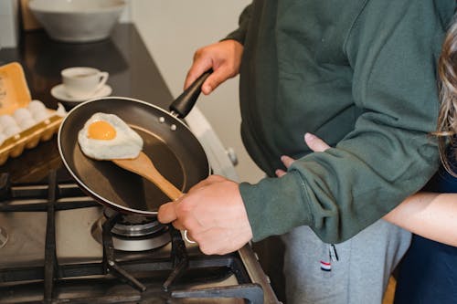 Free Crop anonymous female embarrassing man in casual clothes standing near gas stove with pan in hand and cooking fried egg for diner in kitchen at home Stock Photo