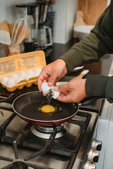 A person cracking an egg into a pan on a stove, preparing a breakfast dish in a cozy home kitchen.