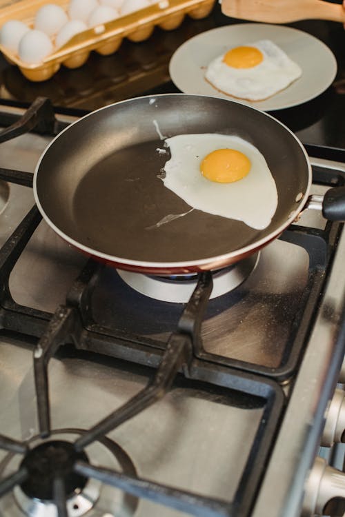 From above of egg frying in pan on gas stove and placed near ready fried eggs on plate in kitchen