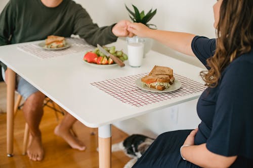 Free Crop anonymous happy couple in casual clothes holding hands above table with sandwiches and plate with fruits before breakfast at home Stock Photo