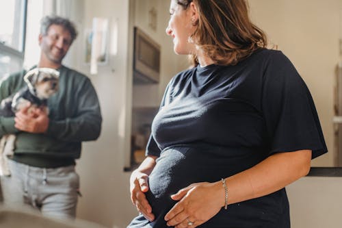 Crop positive pregnant wife touching belly and looking at smiling husband with dog in hands in cozy home in daytime