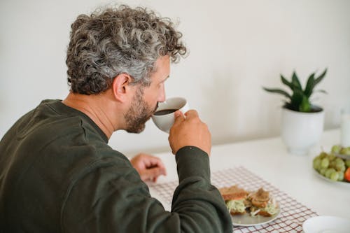 Thoughtful male with beard and curly gray hair in casual outfit drinking coffee while having sandwich for breakfast