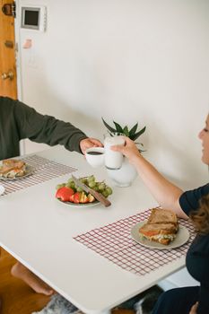 Crop anonymous female and male drinking milk and coffee while having breakfast together in kitchen at home