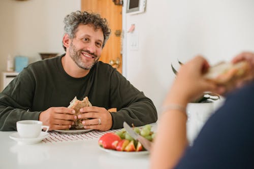Smiling middle aged guy with beard and gray hair in casual wear eating toast while having breakfast with wife in kitchen