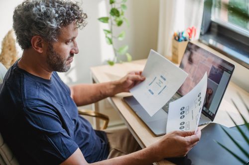 Free Focused adult male with beard and gray curly hair in casual clothes sitting at table with laptop open while working with papers Stock Photo