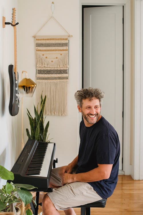 Adult cheerful bearded man with curly gray hair sitting on chair near piano and looking away