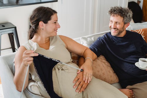 Young pregnant female knitting on sofa while bearded husband touching belly and having morning coffee