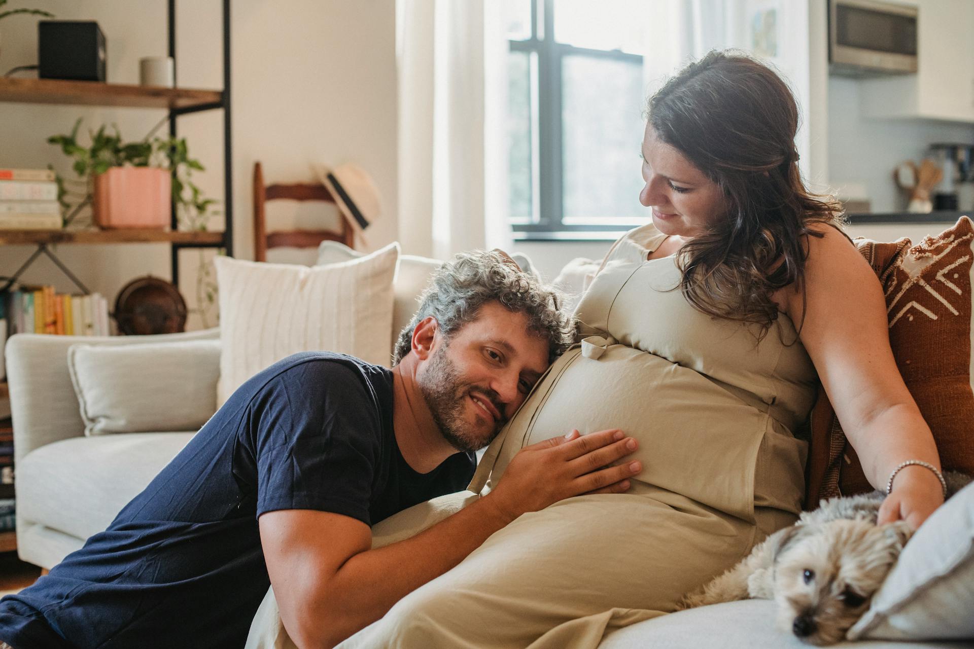 Happy young pregnant female caressing dog while bearded husband cuddling to tummy with cheek