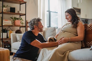 Side view of smiling adult man with beard and gray curly hair looking at happy pregnant wife stroking adorable dog and touching tummy