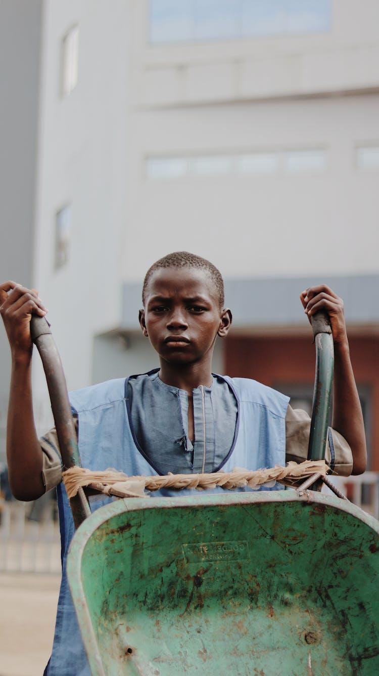 Pensive Black Child With Wheelbarrow By Modern Building