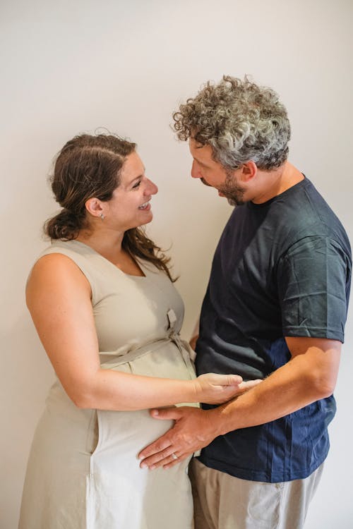 Side view of bearded man touching pregnant tummy of smiling wife in casual dress while standing and talking against white background