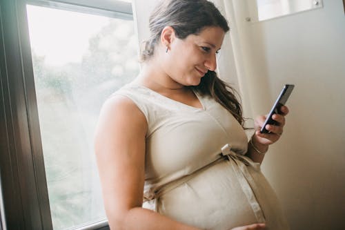 Free Adult pregnant lady checking notifications on smartphone while standing near window in room in daytime Stock Photo