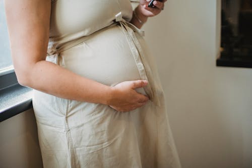 Free Crop unrecognizable pregnant female standing near window and checking notification on cellphone in room Stock Photo