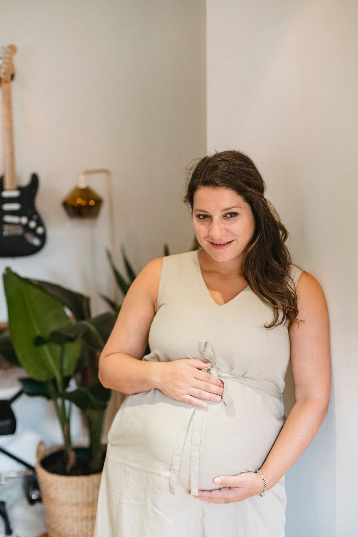 Smiling pregnant woman standing indoors while holding her baby bump, with her face clearly visible